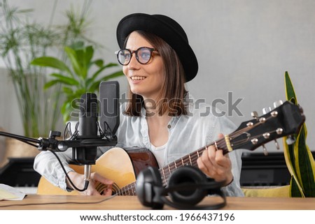 podcast, music audio content creation. beautiful European woman podcaster in a hat with a guitar or ukulele, radio host recording podcast or content, song and music in a recording studio