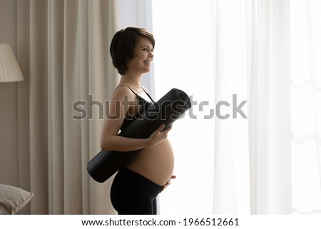 Happy active young pregnant woman wear sportswear hold yoga mat ready for morning gymnastics or pilates. Smiling sporty female follow healthy lifestyle during pregnancy, do exercise training.
