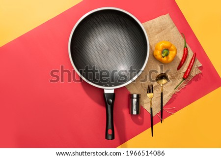 Flat lay of cooking equipment frying pan with chili, salt, tomato, pepper, and paprika with spoon and fork in yellow and red background