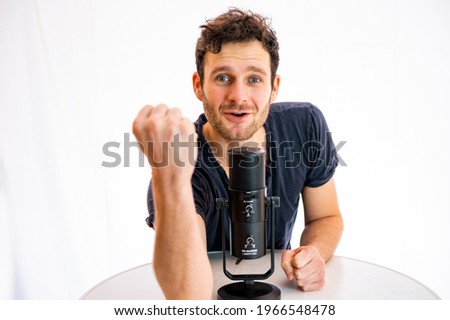 Young man with dark hair and blue eyes in front of a white studio background speaking into a microphone