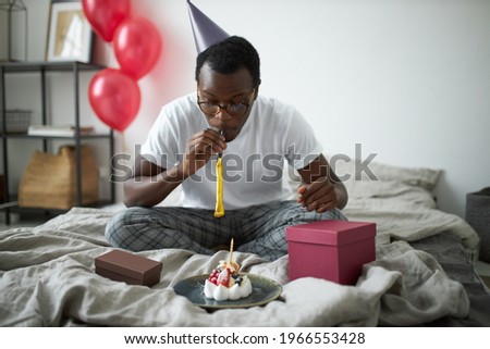 Special occasion, celebration and holidays. Indoor image of happy young dark skinned male wearing glasses and cone hat having fun in bedroom, celebrating his 30th birthday, blowing party horn