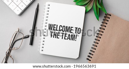 welcome to the team is written in a white notebook with calculator, craft colored notepad, plant, black marker and glasses.