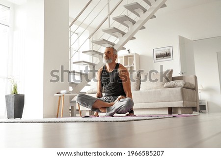 Strong older mid aged tattooed yogi man doing yoga exercises at home.Fit healthy mature senior tattooed male meditating relaxing lifting himself over mat sitting in yoga pose in living room at home. Royalty-Free Stock Photo #1966585207