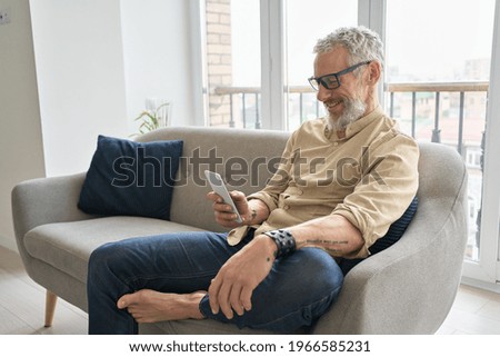 Happy older middle aged man using apps on phone relaxing sitting on couch at home. Smiling senior hipster with tattoos wearing glasses holding cellphone device ordering delivery online or texting. Royalty-Free Stock Photo #1966585231