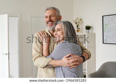 Happy senior adult mature classy couple hugging, bonding, thinking of good future. Carefree cheerful mid age old husband embracing wife looking away dreaming, enjoying wellbeing and love in new house. Royalty-Free Stock Photo #1966585285