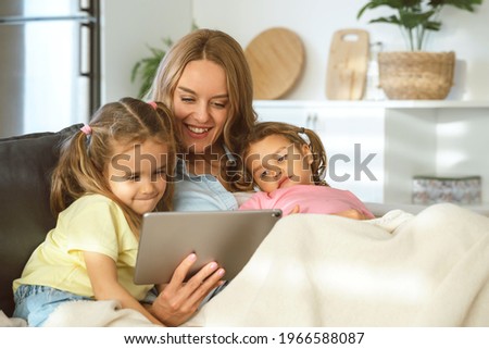 Happy family, young loving mother and two cute little girls sisters having video call with relatives father or grandparents on digital tablet while relaxing on sofa at home. Leisure time with kids