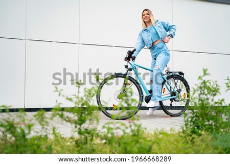 Cute young blonde woman sitting on bicycle and smiling, city ride, bright exterior of modern building in background. Copy space 