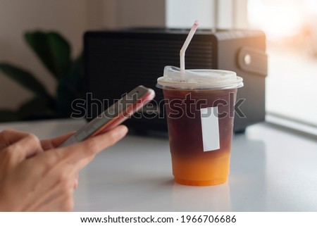A plastic cup of espresso coffee shot with orange juice on mirror table with women hand holding phone