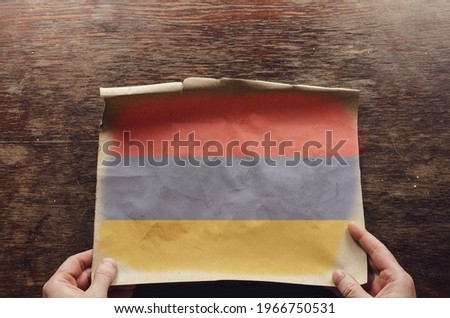 An old, shabby sheet of paper with the Armenian flag on it lies on the scratched table. A man's hands press the unfolded sheet of parchment against the brown wooden table with the Armenian tricolor. 