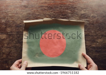 On the scratched table lies an old, tattered piece of paper with a picture of the Bangladeshi flag on it. The man's hands press the unfolded piece of parchment against the brown wooden table. 