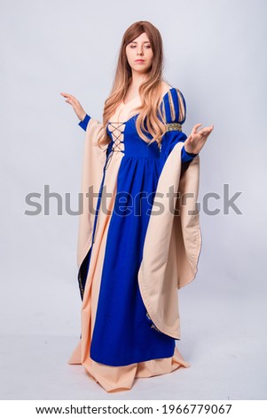 A full-length portrait of an attractive woman with long hair in a medieval, fantasy, blue and beige dress with long, large sleeves, posing isolated on a white background.