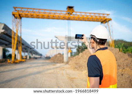 Smart Asian worker man or male civil engineer with protective safety helmet and reflective vest using using smartphone for taking photo at construction site.