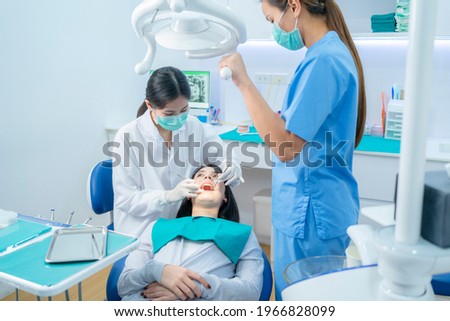 Asian female dentist adjust dental surgical light then starts checking or examining tooth of young girl patient lying on dental chair. Dental assistant support by handing instruments at dental clinic. Royalty-Free Stock Photo #1966828099
