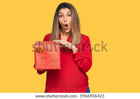 Beautiful brunette woman holding gift scared and amazed with open mouth for surprise, disbelief face 