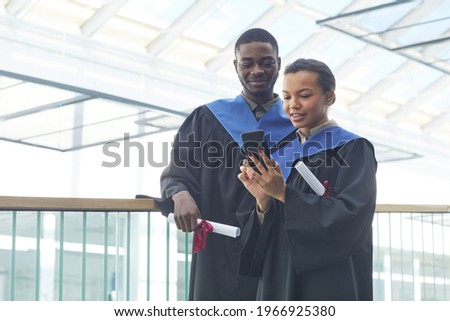 Waist up portrait of young African-American couple wearing graduation gowns while taking selfie indoors in modern university interior, copy space