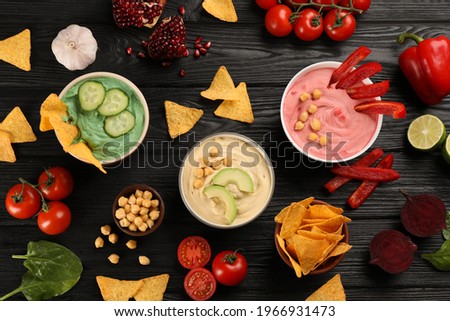 Different kinds of tasty hummus served with nachos on black wooden table, flat lay