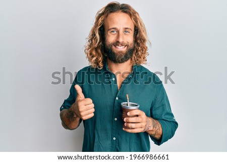 Handsome man with beard and long hair drinking mate infusion smiling happy and positive, thumb up doing excellent and approval sign 