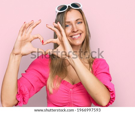 Beautiful caucasian woman wearing sunglasses and summer style smiling in love showing heart symbol and shape with hands. romantic concept. 