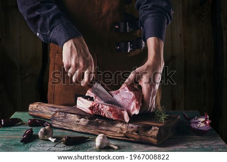 A guy in a leather apron is slicing raw meat. The butcher cuts the pork ribs. Meat with bone on a wooden cutting board. Royalty-Free Stock Photo #1967008822