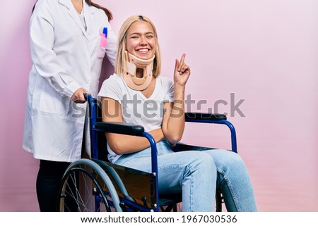 Beautiful blonde woman sitting on wheelchair with collar neck with a big smile on face, pointing with hand and finger to the side looking at the camera. 