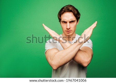 Young man looks seriously and shows not allowed, isolated on green background Royalty-Free Stock Photo #1967041006