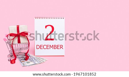 2nd day of december. A gift box in a shopping trolley, dollars and a calendar with the date of 2 december on a pink background. Winter month, day of the year concept.
