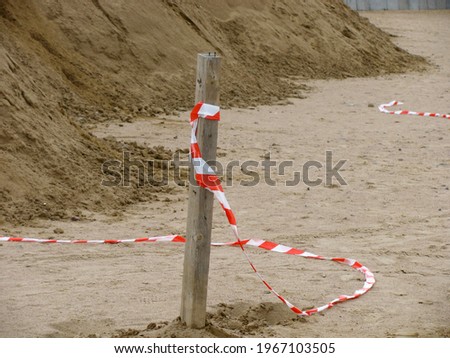 A red and white signal tape is tied to a wooden pole. The ribbon blocks the way to the beach. Ribbon on the sand background.