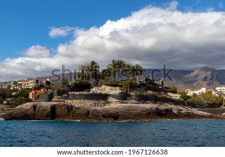 Castle on rocky island with palms and blurred volcanic mountains. Ocean coastline. Blue sky and white clouds.