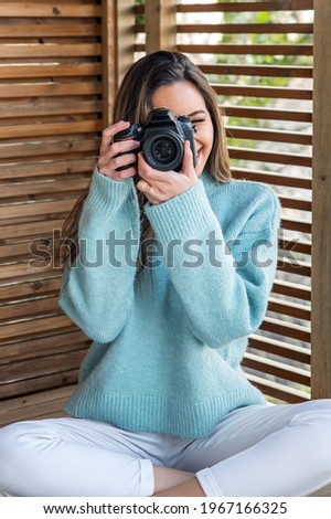 Full body of smiling young female photographer in casual outfit sitting on wooden terrace and taking pictures with photo camera while enjoying sunny weekend