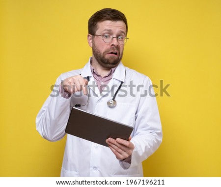 Surprised doctor holds a tablet in hands and excitedly points finger at the screen. Yellow background.