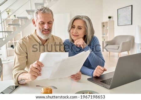 Middle aged senior old couple holding documents reading paper bills paying bank loan online, calculating pension fees, payments, taxes, planning family retirement money finances using laptop at home. Royalty-Free Stock Photo #1967198653