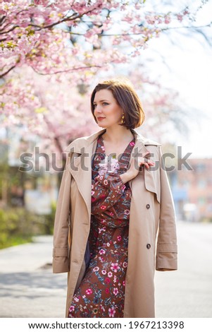 Portrait of a gentle woman against the background of sakura flowers. Walk in the blossoming sakura garden. Young stylish woman standing in sakura park and enjoying beauty