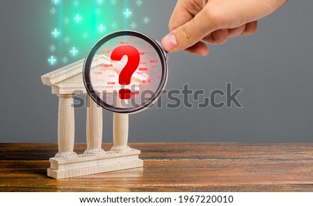 A hand with a magnifying glass inspects bank and finds financial violations. Audit and accounting. Money laundering, sanctions evasion. Insolvency, misleading. Low bank liquidity, risk of bankruptcy. Royalty-Free Stock Photo #1967220010