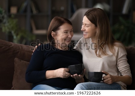 Smiling grownup Caucasian female kid child relax rest on couch with mature 60s mom drink coffee tea together. Happy two generations of women enjoy family reunion at home. Relation concept. Royalty-Free Stock Photo #1967222626