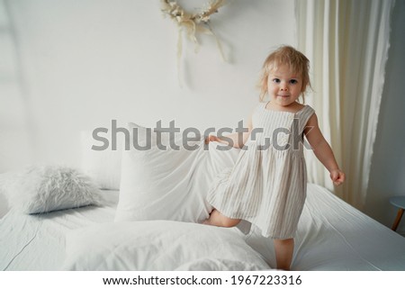 Small child blonde European appearance girl playing in bed. White bed linen. Beautiful children's white stylish modern dress. Time to rest, get ready for bed.