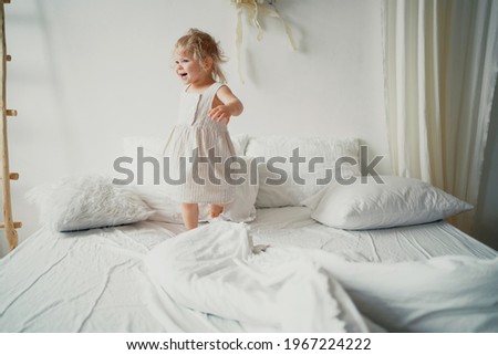 Baby blonde European-looking girl playing in bed. Beautiful children's white stylish modern dress. White bed linen. Time to rest, get ready for bed. Good morning in the bedroom.