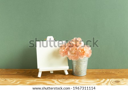 Easel and blank canvas with carnation flowers on wooden table. green background