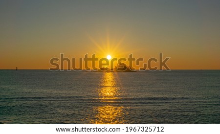 The sun is rising up over an island on the sea. Sunlight shines like a star and sky glows red and gold colors and ocean surface reflects sunshine. Beautiful nature scene took with aerial drone picture