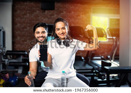 Sporty Indian asian man and woman resting between exercises and using smartphone at gym, presenting something or taking selfie picture
