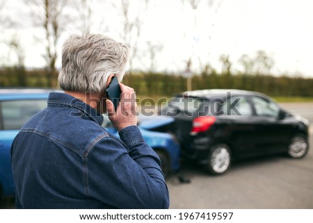 Rear view of senior male driver standing by damaged car after traffic accident reporting incident to insurance company using mobile phone Royalty-Free Stock Photo #1967419597