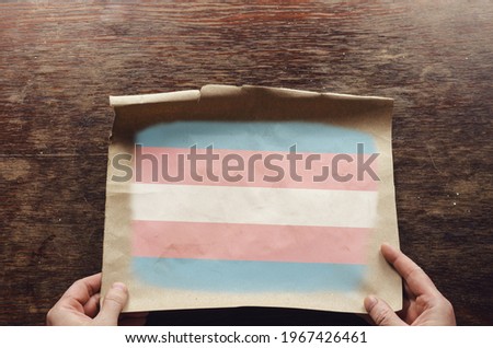 On the scratched table lies an old, tattered piece of paper with a picture of the Transgender pride flag. The man's hands press the unfolded piece of parchment against the brown wooden table