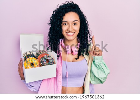 Young hispanic woman with curly hair holding tasty colorful doughnuts box smiling with an idea or question pointing finger with happy face, number one 