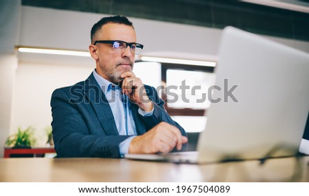 Skilled businessman in optical eyewear for vision protection checking online exchange on modern netbook technology using office internet during work day, Caucasian male expert reading financial news
