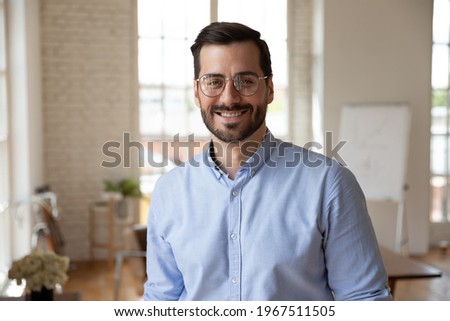 Portrait of smiling young 30s handsome bearded businessman in eyeglasses posing in modern workplace. Happy confident male entrepreneur worker employee looking at camera, professional career concept. Royalty-Free Stock Photo #1967511505