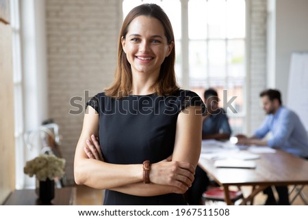 Portrait of confident smiling attractive female team leader in formal dress standing in modern workplace. Happy young 30s businesswoman boss employer partner posing indoors, looking at camera. Royalty-Free Stock Photo #1967511508