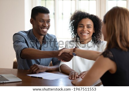 Happy young african american married couple clients shaking hands with financial advisor, discussing contract terms of conditions. Emotional glad millennial diverse spouses celebrating making deal. Royalty-Free Stock Photo #1967511553