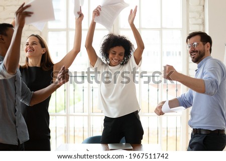 Happy young african american female team leader having fun with multiracial colleagues, dancing to energetic disco music, throwing papers in air, celebrating business success or achievement together. Royalty-Free Stock Photo #1967511742