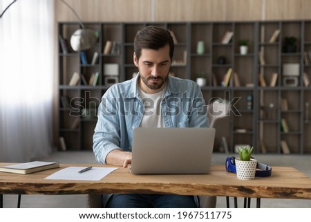 Confident businessman working on laptop online, typing, writing business email or financial report, sitting at wooden desk at home, focused young man searching information, educational course