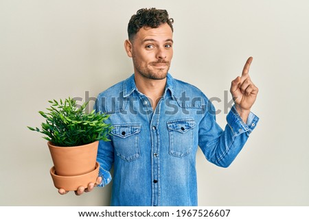 Young hispanic man holding wooden plant pot smiling happy pointing with hand and finger to the side 