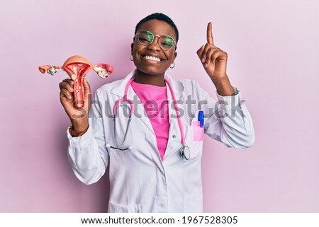 Young african american doctor woman holding anatomical model of female genital organ smiling with an idea or question pointing finger with happy face, number one  Royalty-Free Stock Photo #1967528305